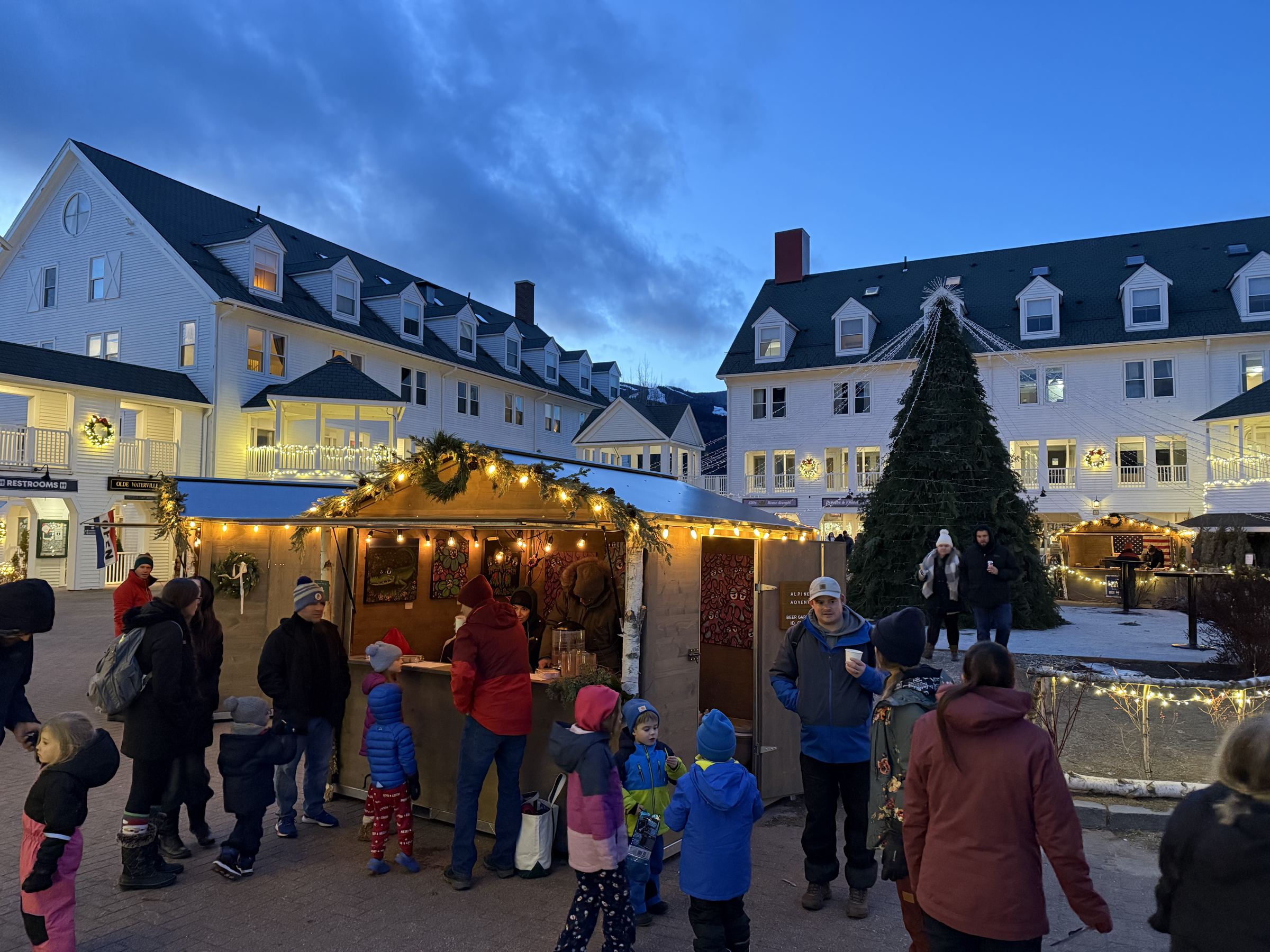Shoppers and families at a lit chalet beside the Christmas tree in Waterville Valley's Town Square at dusk